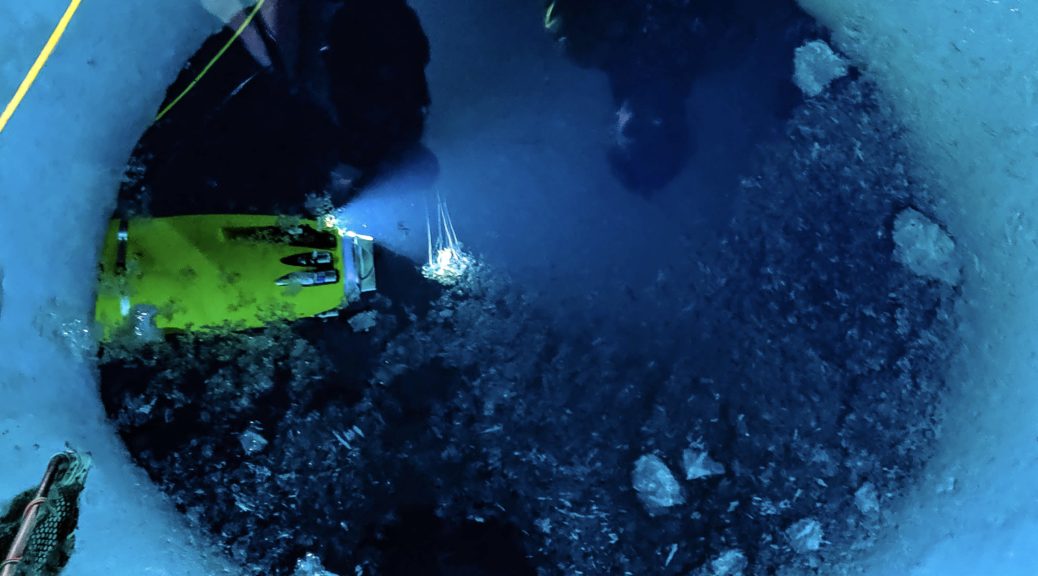 Picture of a hole in ice, with dark ocean beneath the ice. In the water, the yellow Icefin Robot shines light on a small jellyfish near the robot.
