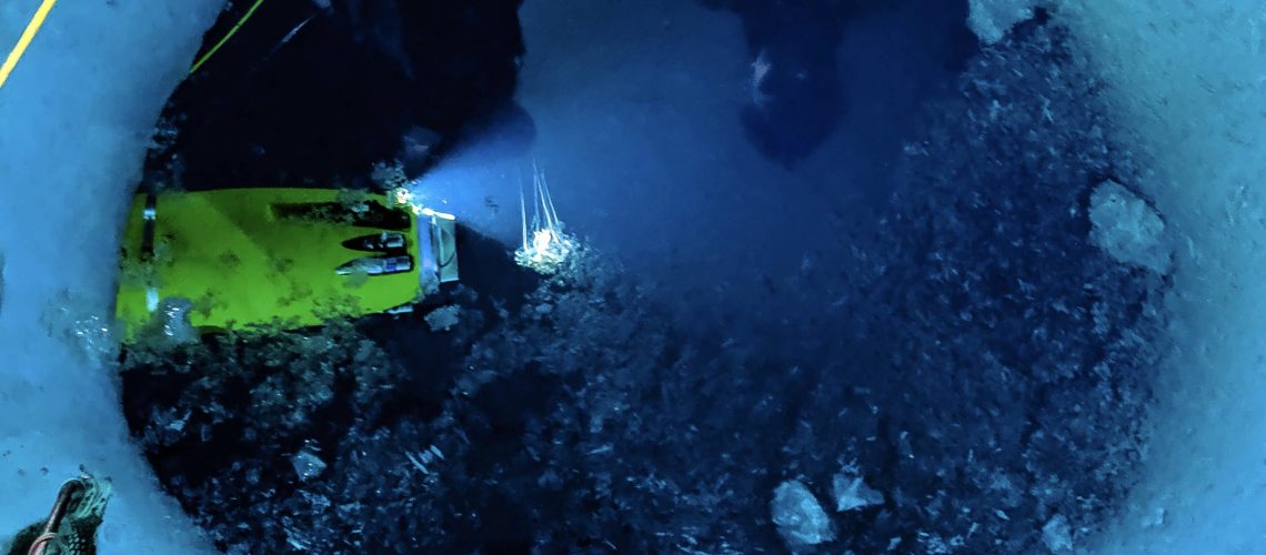 Picture of a hole in ice, with dark ocean beneath the ice. In the water, the yellow Icefin Robot shines light on a small jellyfish near the robot.